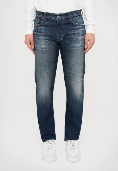 Jean en denim bleu foncé avec une coupe droite, un devant délavé, cinq poches et des détails de couture orange. Associé à des baskets blanches.
