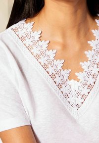 Žena v bílém tričku s V výstřihem a květinovou krajkovou aplikací kolem krku a krátkými rukávy.