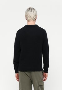 Pulover negru din tricot cu guler rotund, manșete și tiv cu dublu tors, având un finisaj texturat, prezentat din spate. Stilizat cu pantaloni de culoare verde măslin.