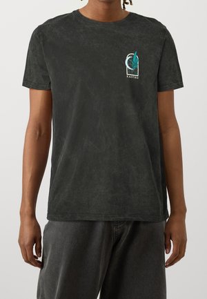 T-shirt noir avec une finition texturée, présentant un imprimé graphique avec des éléments verts et bleus sur la poitrine. Design à manches courtes et encolure ronde.