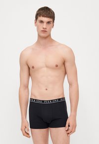 Schwarze Baumwoll-Boxershorts mit elastischem Bund, auf dem das wei�ße Logo "PIER ONE" abgebildet ist. Das Modell steht mit hängenden Armen und hat einen neutralen Gesichtsausdruck.