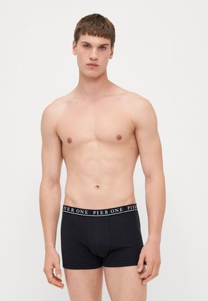 Schwarze Baumwoll-Boxershorts mit elastischem Bund, auf dem das weiße Logo "PIER ONE" abgebildet ist. Das Modell steht mit hängenden Armen und hat einen neutralen Gesichtsausdruck.