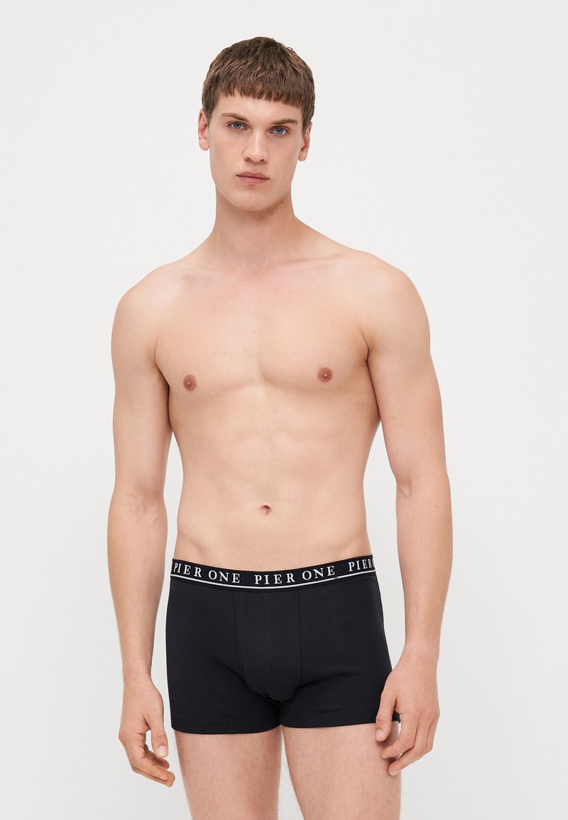 Schwarze Baumwoll-Boxershorts mit elastischem Bund, auf dem das weiße Logo "PIER ONE" abgebildet ist. Das Modell steht mit hängenden Armen und hat einen neutralen Gesichtsausdruck.