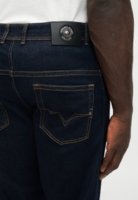 Οπίσθια όψη ενός ατόμου που φοράει σκούρο μπλε τζιν με πορτοκαλί ραφές και μια μαύρη ετικέτα Versace Jeans Couture στη μέση του παντελονιού.