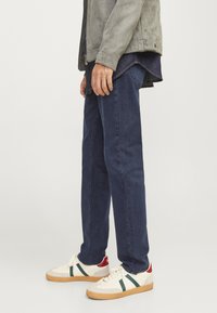 Dark blue denim jeans with straight fit, worn with a light gray suede jacket and white sneakers featuring green and red accents.