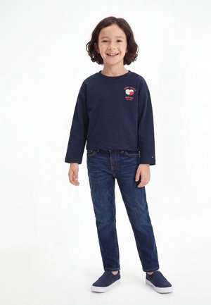 Kind met donker haar tot op de schouders, draagt een marineblauwe trui, donkerblauwe jeans en marineblauwe slip-on schoenen, glimlacht en staat voor een witte achtergrond.