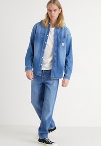 Camicia in denim blu con chiusura a bottoni e tasche, abbinata a pantaloni in denim blu; le caratteristiche includono una vestibilità rilassata e sneakers nere.