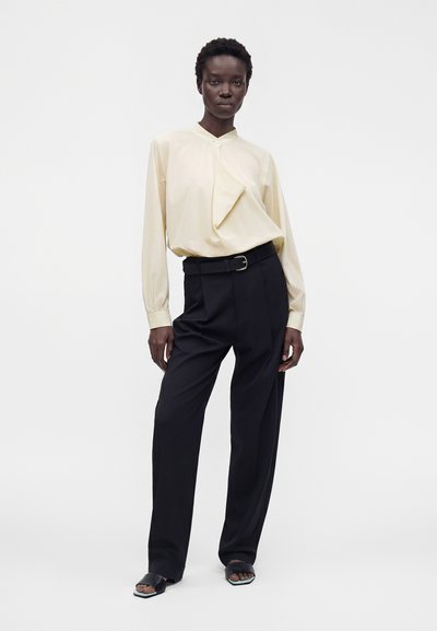 Personne portant un chemisier crème à manches longues avec un détail de pli, un pantalon large noir ceinturé, et des sandales noires à bout ouvert, debout sur fond blanc.