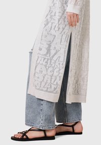 Cardigan lungo e leggero in maglia aperta di colore grigio chiaro con motivi geometrici, abbinato a jeans a gamba larga in denim e sandali neri flat.