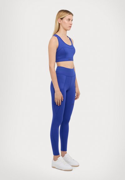 Soutien-gorge de sport bleu et leggings taille haute en tissu extensible. Comprend des détails de couture et un branding discret. Associé à des baskets blanches.