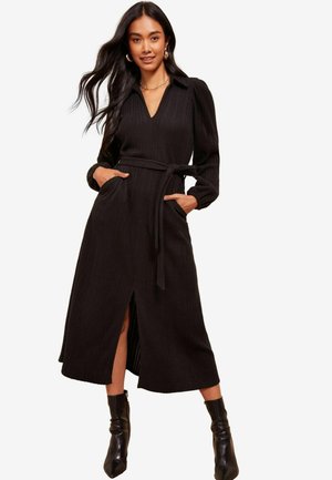 Abito maxi nero a coste con colletto, maniche lunghe, vita con cintura, tasche laterali e spacco frontale. Abbinato a stivali neri alla caviglia.