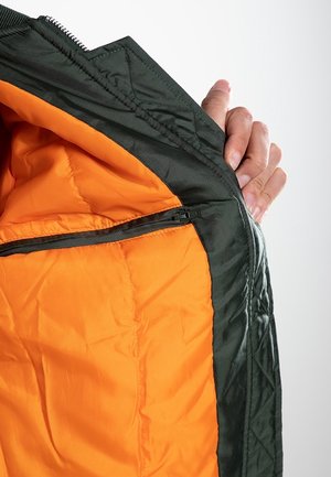 Hand die een open zwart jack vasthoudt om een feloranje doorgestikte voering te tonen met een zwarte ritssluiting langs de binnenrand.