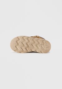 Suela de zapato de goma de color marrón claro con un patrón ondulado, con parte superior de textil gris y una correa decorativa con detalles metálicos.