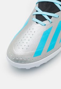 Zapatilla deportiva plateada con acentos turquesas, parte superior texturizada que presenta pequeños bultos y lengüeta de malla negra. Suela blanca con clavos para tracción.