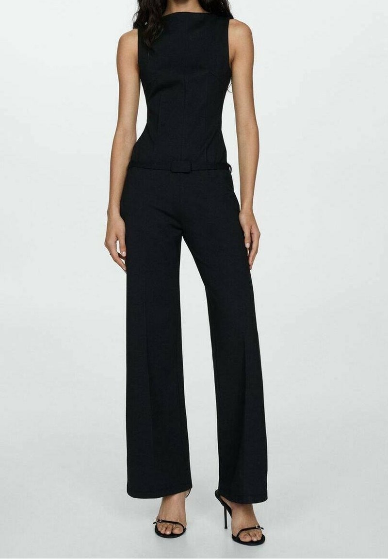 Femme portant une combinaison noire sans manches avec un pantalon à jambes larges et une ceinture assortie, associée à des sandales noires à talons hauts.