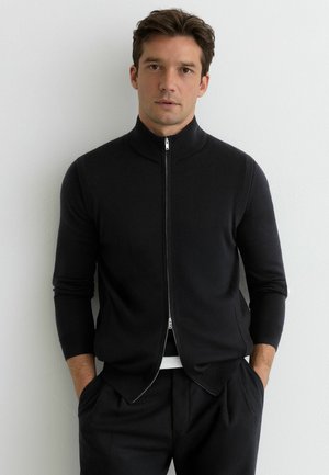 Czarny sweter na zamek z wysokim kołnierzem i długimi rękawami. Wykonany z miękkiej, gładkiej tkaniny, charakteryzuje się opływowym krojem i bocznymi kieszeniami.