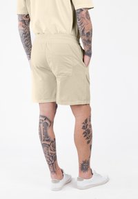 Man in beige shorts en een bijpassend shirt staat met zijn handen in zijn zakken, toont tatoeages op zijn armen en benen, en draagt witte sneakers.