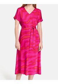 Różowa sukienka o kroju kopertowym z krótkimi rękawami, dekoltem w kształcie litery V oraz wiązanym paskiem w talii. Materiał zdobi falisty wzór w odcieniach różu i pomarańczu.