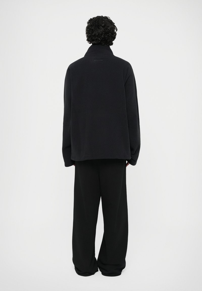 Black, long-sleeve fleece pullover with a high collar, worn over loose-fitting black pants. Smooth texture, minimal design, and no visible fastenings.