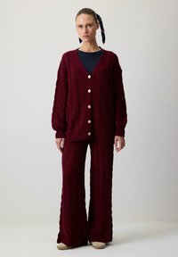 Sieviete, tērpusies bordo krāsas adītā kardigānā ar pogām un saskaņotās platās biksēs, stāvot uz vienkārša gaiša fona.