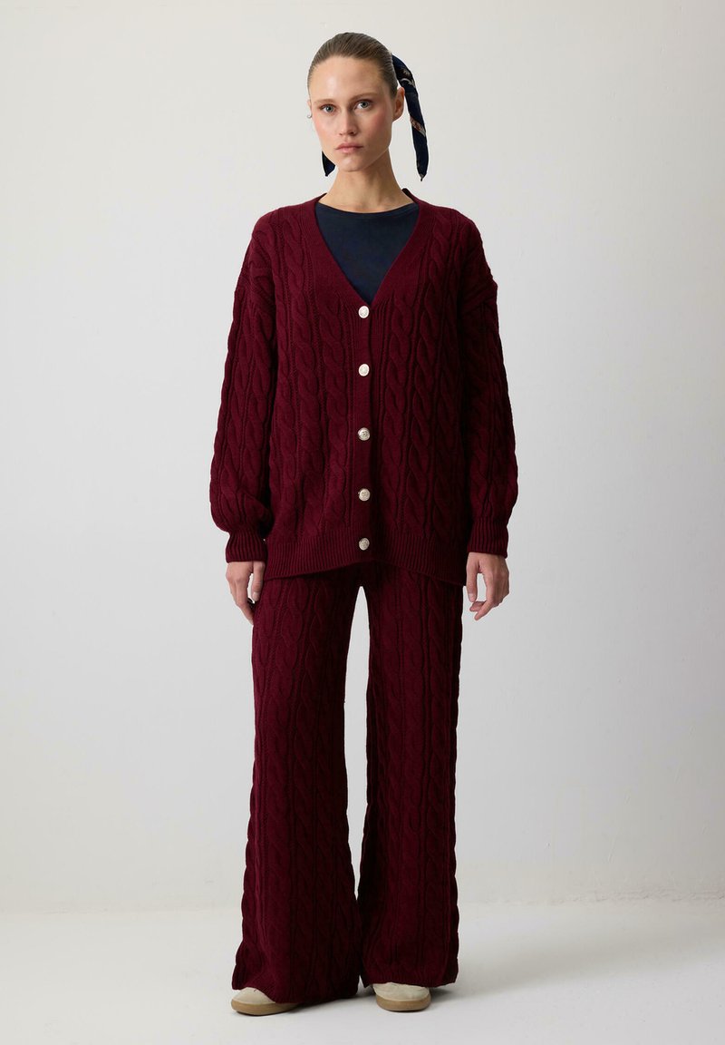Sieviete, tērpusies bordo krāsas adītā kardigānā ar pogām un saskaņotās platās biksēs, stāvot uz vienkārša gaiša fona.