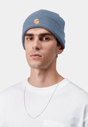 Jasnoniebieska, prążkowana czapka beanie z małym żółtym logo. Noszona na białej bluzie z widoczną srebrną łańcuszkiem.