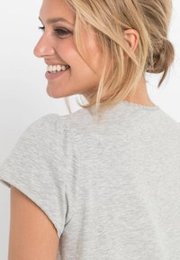 Maglietta grigia a maniche corte con un design semplice, texture morbida e scollatura ondulata, vista da dietro con un leggero volume sulle spalle.