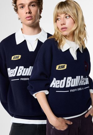 Junger Mann und Frau tragen dunkelblaue Red Bull Racing Pullover mit weißen Kragen und stehen eng nebeneinander vor einem einfarbig hellen Hintergrund.