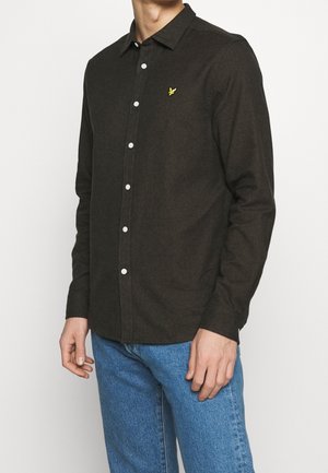 Homme portant une chemise marron foncé boutonnée avec un petit logo d'oiseau jaune sur la poitrine et un jean bleu, debout devant un fond uni.