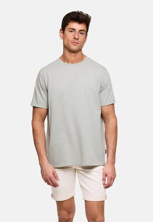 Joven de pie que lleva una camiseta de manga corta gris claro y pantalones cortos beige remangados, sobre un fondo blanco simple.