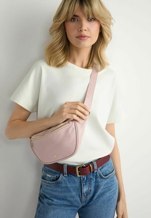 Blondynka w białej bluzce i dżinsach nosi jasnoróżową torbę na ramię z złotym zamkiem oraz bordowy pasek ze złotą klamrą.