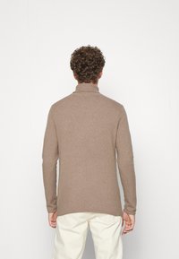 Man med lockigt hår som bär en beige polotröja och ljusa byxor, stående vänd mot en vit vägg.