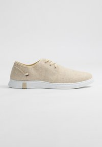 Zapatillas de tela beige con superficie texturizada, suela de goma blanca y un detalle sutil de marca en el lateral. Diseño con cordones y punta redondeada.