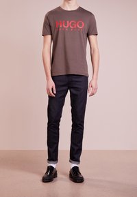 Braunes Baumwoll-T-Shirt mit rotem "HUGO BOSS"-Schriftzug, kombiniert mit dunkelblauen Jeans. Das Modell trägt schwarze Schuhe mit weißen Socken und steht auf einem hellen Boden.
