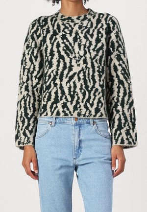 Mujer con un suéter de patrón abstracto en negro y beige y jeans de cintura alta color azul claro contra un fondo liso.