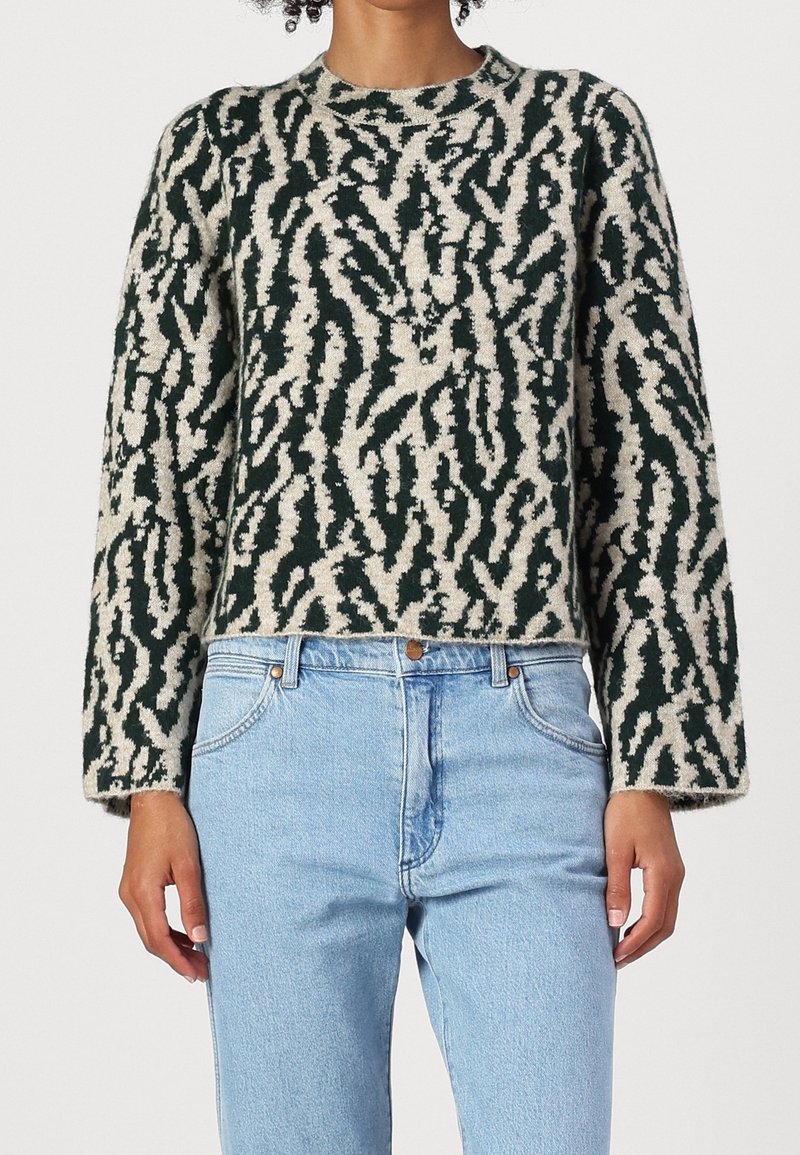 Femme portant un pull à motifs abstraits noir et beige avec un jean taille haute bleu clair devant un fond uni.