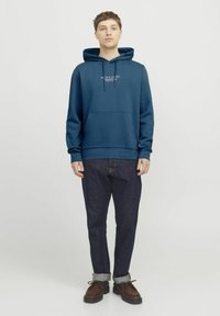Felpa blu con cappuccio e cordini, abbinata a jeans blu scuro e scarpe stringate marroni. Tessuto morbido, vestibilità casual.