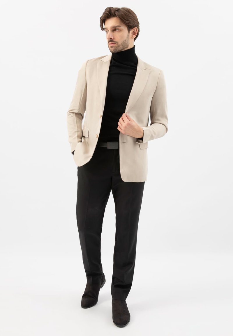 Uomo che indossa un blazer beige, dolcevita nero, pantaloni neri e scarpe nere, in piedi con una mano che sistema il blazer, guardando di lato.