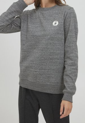 Personne portant un pull gris chiné à manches longues avec un petit logo blanc circulaire sur la partie supérieure de la poitrine et un pantalon gris foncé.