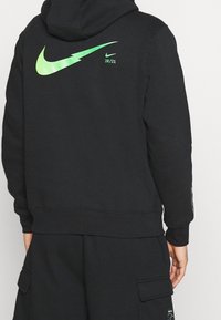 Svart huvtröja i mjukt tyg, med en stor grön Nike-logotyp på ryggen och texten "20/21". Ribbad mudd på ärmsluten och midja, samt detaljerad känguruficka.