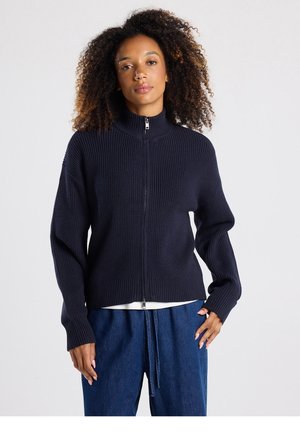 Marineblauwe gebreide trui met een hoge kraag, volledige ritssluiting, geribbelde textuur en een losse pasvorm. Heeft brede mouwen en een cropped zoom.