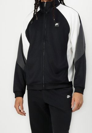Personne portant une veste zippée Nike Air noire, blanche et grise et un pantalon de jogging Nike noir, debout devant un fond uni.