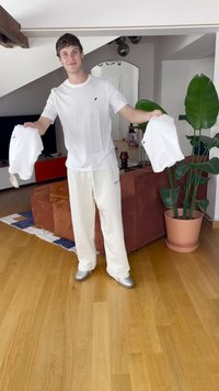 White short-sleeve shirt, cream sweatpants, and silver shoes. Person holding two folded white shirts in an indoor setting with wooden floor.