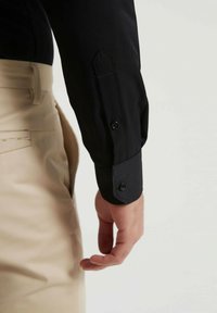 Chemise noire à manches longues avec poignet boutonné, présentant un design épuré et une texture de tissu, associée à un pantalon beige avec des détails de poche visibles.