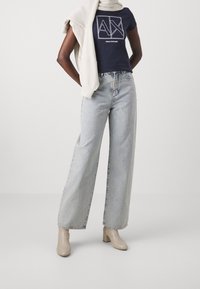 Námořnické tričko s bílým logem "Armani Exchange", v kombinaci se světle modrými širokými džíny a béžovými kotníkovými botami na podpatku. Lehký šátek přehozený přes ramena.