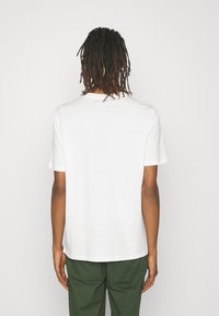 Witte katoenen T-shirt met korte mouwen en een ronde halslijn, met een rechte zoom. G gedragen met groene broek, gezien vanaf de achterkant.
