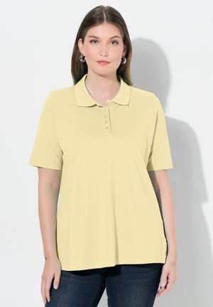 Kobieta o prostych brązowych włosach, ubrana w jasnożółtą koszulkę polo z krótkim rękawem i ciemne dżinsy, stojąca na białym tle.