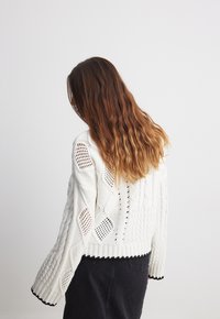 Gruby biały sweter dziergany z teksturowanymi wzorami i otwartymi, diamentowatymi wycięciami. Mankiety i dół z prążkowanego materiału z czarną lamówką.