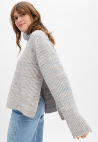 Jonge vrouw in een lichtgrijze geribbelde turtleneck trui en blauwe jeans, zijwaarts staand met iets uitgestrekte armen tegen een eenvoudige achtergrond.