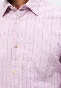 Ανοιχτό ροζ πουκάμισο με λεπτές κάθετες μαύρες ρίγες και τσέπη στο στήθος, που φοριέται από άτομο με καστανό δέρμα.